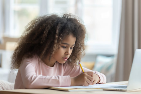 African American little girl with curly hair sitting at the table, writing and doing homeworkの素材