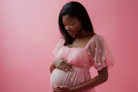 9 month pregnant African American woman wears pink dress touching belly against pink backgroundの素材