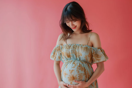 9 month pregnant Asian woman wears flower printed dress touching her belly against pink backgroundの素材