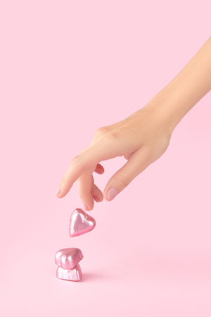Beautiful well-groomed female hand with pink matte nails and heart-shaped candies on a pink...