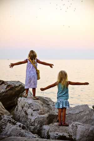 Little blonde girls in summer dress standing on rocks and staring at ocean with open armsの写真素材