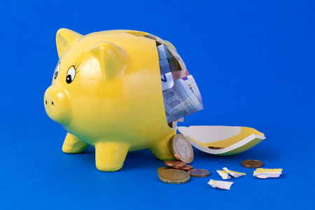 Yellow broken piggy bank containing euro bills and coins on blue backgroundの写真素材