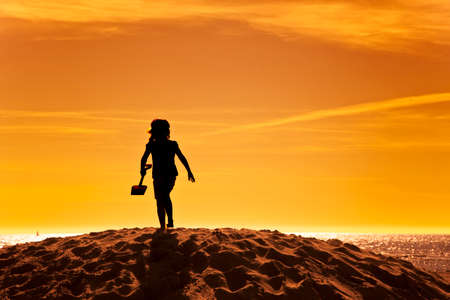 silhouette of little girl with shovel on a sand hill at the beach in sunsetの写真素材