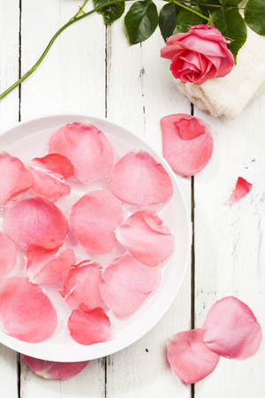 spa concept, pink rose petals in bowl of water with towel and flower on white wood background, high angle viewの写真素材
