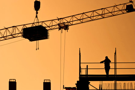 Silhouette of worker standing on scaffolding and load hanging on crane at construction site in...