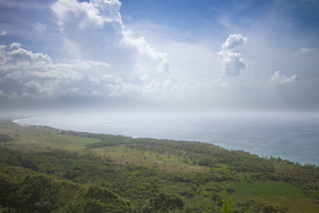 Lush green fields stretch out toward a vast, misty ocean under a dramatic sky filled with billowing...