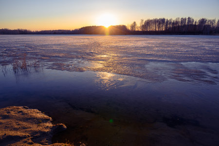 Colorful sunset with sun beams on the shore of a lake in early spring. Reeds and trees in the...