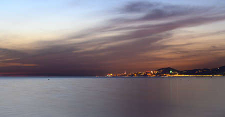 Plain water surface of the Black Sea. Dark clouds are colored with the set sun. Lights of the city Adler in the background. The beauty of sea sunsets.の写真素材