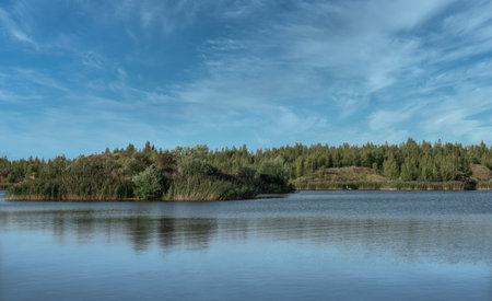 A lake with a plane surface and reflections against the blue sky and white clouds. Originated from an old quarry flooded, groups of islands overgrown with reeds. Beauty in nature, landscapes, scenic.の写真素材