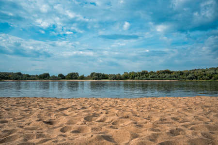 Sandy bank of a river with reflections on its surface and trees in the distance against a grey cloudy sky. Autumn landscape with rural views.の写真素材