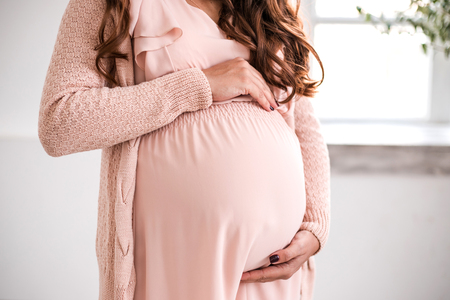 Beautiful pregnant woman is holding belly close-up. Light from the window holding her. Concept of waiting baby and hapiness of of motherhood.の写真素材