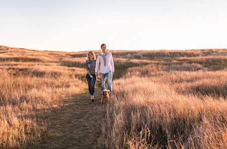 Happy young couple is walking with dog outdoors. Sunset and beautiful gold light.の写真素材