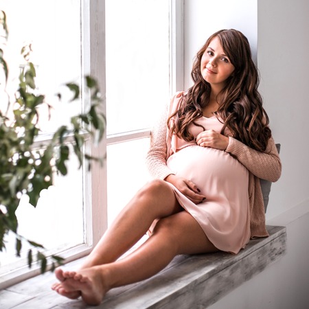 Beautiful smiling pregnant woman is holding belly and looking at camera. Light from the window holding her. Concept of waiting baby and hapiness of of motherhood.の写真素材