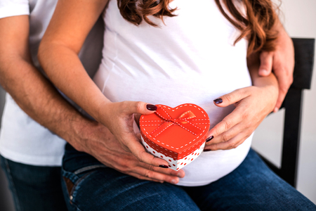 Happy parents hug and hold pregnant belly. Woman hold a gift from her husband. They are happy to celebrate Valentines Day together.の写真素材