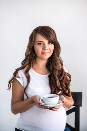 Happy pregnant woman with cup of tea sitting on the chair near the window. Concept of healthcare and maternity.の写真素材
