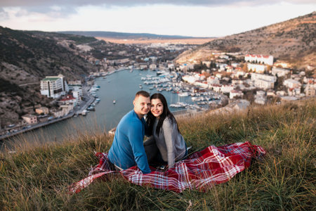 Happy couple seating on the hill near Black sea. Amazing view of the bay. Young attractive travellers smiling and hugging.の写真素材