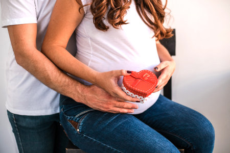 Happy parents hug and hold pregnant belly. Woman hold a gift from her husband. They are happy to...