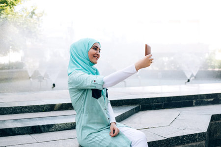 Beautiful young muslim woman making selfie by smartphone outdoor. Woman wearing hijab using mobile...
