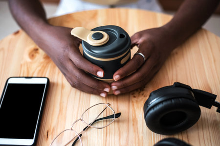 Pretty young black girl listening to music on smartphone at cafe. She drinks fresh hot coffee at...