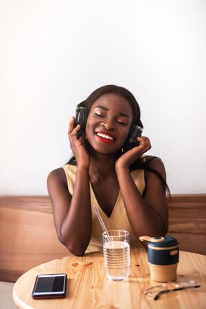 Smiling black girl listening to music on smartphone at cafe. She drinks coffee at reusable cup and singing the song.の写真素材