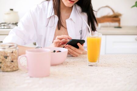Young beautiful woman using mobile phone while healthy breakfast in light modern kitchen.の写真素材