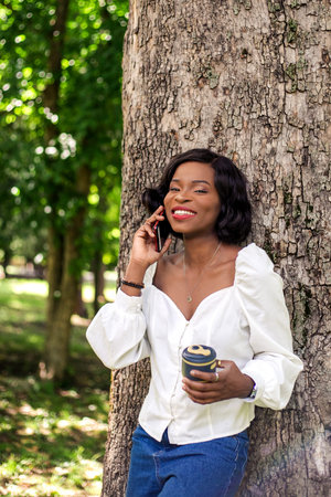 Beautiful african woman talking by phone standing near tree in the park with reusable cup of coffee.