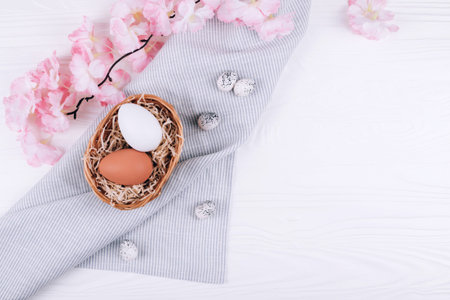 Easter eggs in the nest on textile napkin with sakura blossom on white wooden background.