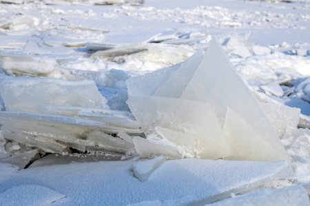 Jagged ice crystals rest on a snow-covered shore, capturing the cold, quiet mood of winter. A stark,...