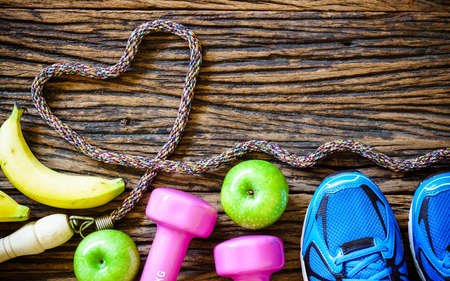 Fitness workout love and healthy fruit eating concept - Top view of bananas, green apples, sport shoes, dumbbells and jump rope in heart shape on wooden backgroundの写真素材