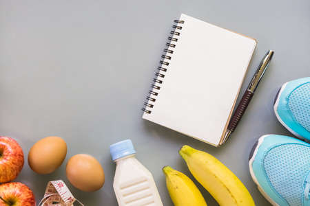 Dieting, healthy and active lifestyles Concept, sport shoes, tape measure, bottle of milk, apples, bananas and blank notebook on grey background. top view with blank copy spaceの写真素材
