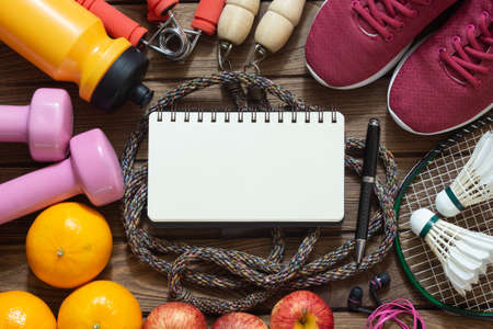 Fitness and healthy active lifestyle dieting background with blank space notebook for your text. Dumbbell, jump ropes, apples, badminton, oranges and blank notebook on wood background. Top view with copy space.の写真素材