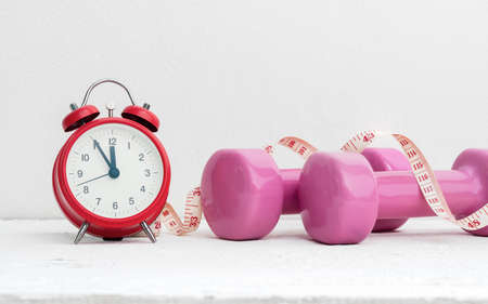 Time to work out, healthy lifestyle and diet concept. Pink dumbbells, alarm clock, and measuring tape on white wood with copy space.の写真素材