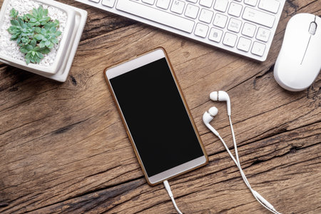 Top view of blank smartphone,  earphones, keyboard and mouse on grunge wooden desktop. Flat lay mock...