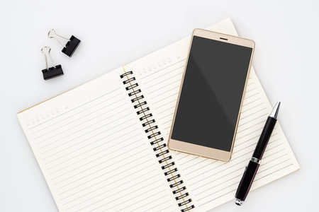 Mock up blank screen of smartphone on notebook with pen  on white background. Flat lay top view with...