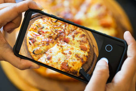 Food blogger taking picture of cooked pizza. Woman's hands with smartphone takes picture of fresh baked hot self-made pizza.  Photography blogging workshop concept.の写真素材