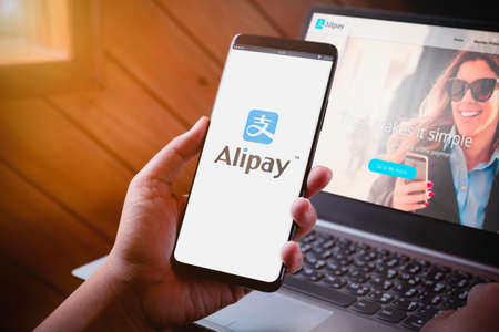 Bangkok, Thailand - August 5, 2019: Hands holding Smartphone with Alipay logo on screen and Alipay website on laptop background. Alipay is mobile and online payment platform, established by Alibaba group.のeditorial素材