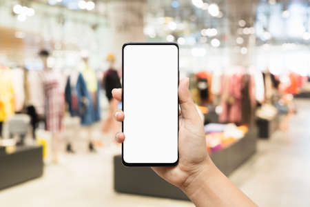 Mockup mobile phone image of woman hand holding smart phone with blank white screen for your advertisement on blurred abstract background of women clothing fashion shop.の写真素材