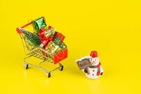 Shopping cart with christmas tree and miniature gift boxes with snowman doll on yellow background. Christmas and New Year shopping time background concept.の写真素材