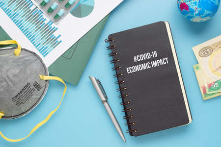 COVID-19 or Corona virus Economic IMPACT background concept. Mockup Notebook for Covid 19 Economic impact anlysis with busines graphs sheets, N95 facial masks, bank notes on blue working desk.の写真素材