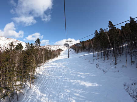 On a sunny winter day in a ski resort, the lift goes up, carries people, top view from a chair lift on a snowy background. A cable car that transports skiers and snowboarders on a winter slope.の写真素材