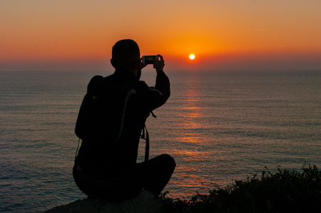Sunset in front of the sea in golden hour with silhouette of hiker taking a picture with his mobile phoneの写真素材