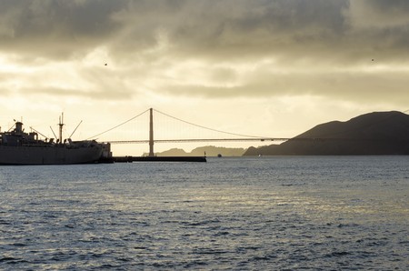 A side view of the famous San Francisco Golden Gate Bridge in California, United States at sunset. A profile view from the Bay, some seagulls flying and the suspended bridge is a silhouette against the golden sky.の写真素材