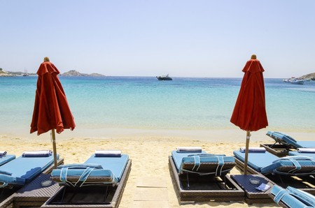 Sun beds with couchins, umbrellas, golden sand and the blue sea in Mykonos, Greece. A greek island summer holiday scene at the Psarou beach with a boat in the crystal clear water.の写真素材