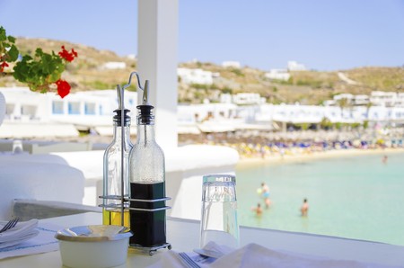 A table set with plates, glasses, olive oil and balsamic vinegar on a beach bar restaurant and a view of the blue sea in Mykonos, Greece. A greek island scene at the beach ready for lunch.の素材 [FY31051645972]