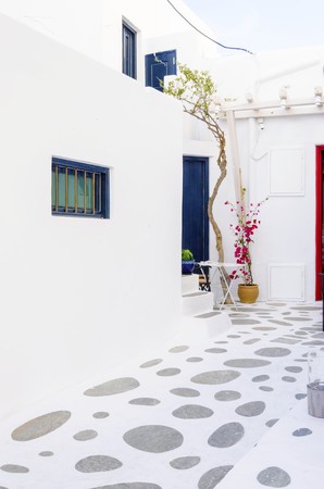 A very traditional alley view of the architecture in Chora, on the greek island Mykonos, Greece. A blue door and windows, a red bougainvillea and flowers outside a whitewashed house and cobble paved street.の写真素材