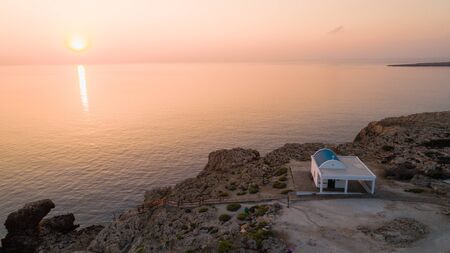 Aerial view of coastline sunset and landmark white washed chapel Agioi Anargyroi, at Cavo Greco Protaras, Famagusta, Cyprus from above. Bird's eye view of tourist attraction cliff rock Ayioi Anargiroi church, caves, beach at sunrise in Ammochostos district.の写真素材