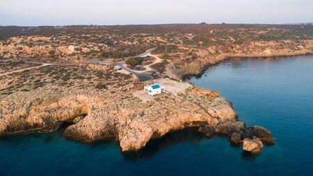 Aerial view of coastline sunset and landmark white washed chapel Agioi Anargyroi, at Cavo Greco Protaras, Famagusta, Cyprus from above. Bird's eye view of tourist attraction cliff rock Ayioi Anargiroi church, caves, beach at sunrise in Ammochostos district.の写真素材