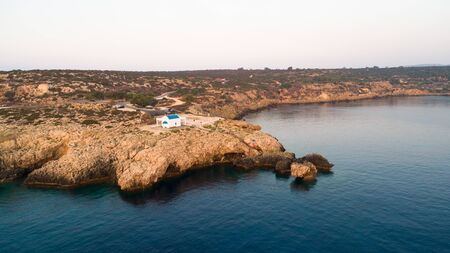Aerial view of coastline sunset and landmark white washed chapel Agioi Anargyroi, at Cavo Greco Protaras, Famagusta, Cyprus from above. Bird's eye view of tourist attraction cliff rock Ayioi Anargiroi church, caves, beach at sunrise in Ammochostos district.の写真素材