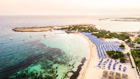 Aerial bird's eye view of famous Makronissos beach coastline, Ayia Napa, Famagusta, Cyprus. The landmark tourist attraction Makronisos bay at sunset with golden sand, sunbeds, sea restaurants in Agia Napa on summer holidays, from above.の写真素材