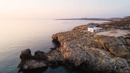 Aerial view of coastline sunset and landmark white washed chapel Agioi Anargyroi, at Cavo Greco Protaras, Famagusta, Cyprus from above. Bird's eye view of tourist attraction cliff rock Ayioi Anargiroi church, caves, beach at sunrise in Ammochostos district.の写真素材